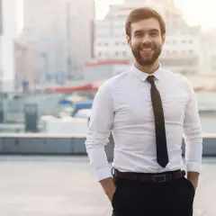Uśmiechnięty mężczyzna w białej koszuli i czarnych spodniach, z rękami w kieszeniach, na tle miejskiego krajobrazu.