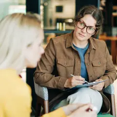 Psycholog a psychoterapeuta w okularach uważnie słucha pacjentki, notując coś na kartce.