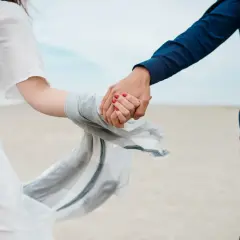 Dłonie splecione na plaży, symbol trwałych związków. Kobieta w zwiewnej białej sukience, mężczyzna w granatowej koszuli.