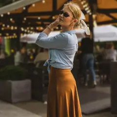 Stylowa kobieta w letniej stylizacji, pokazuje jak zawiązać koszulę. W tle ogródek kawiarniany.