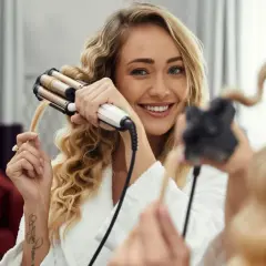 Uśmiechnięta blondynka kręci włosy lokówką trójramienną.