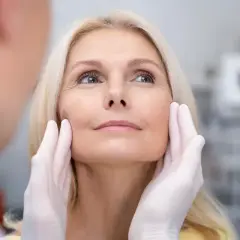 Kobieta z blond włosami poddawana ocenie przed zabiegiem, który może być liftingiem twarzy. Dłonie w rękawiczkach delikatnie dotykają jej policzków.