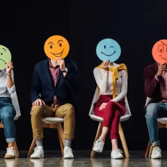 Cztery osoby siedzą na krzesłach, zasłaniając twarze okrągłymi kartkami z różnymi emotikonami. Widać, ile jest emocji: smutek, radość, mrugnięcie i złość.