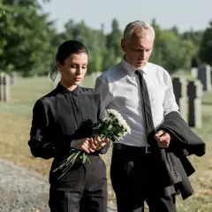 Para pogrzebowa w letni dzień: kobieta w czarnej sukience i mężczyzna w białej koszuli z krawatem, niosący marynarkę i kwiaty.