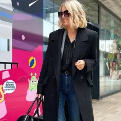Elegancka stylizacja na imprezę: czarny płaszcz, jeansy i stylowe dodatki. Kobieta z blond włosami i okularami przeciwsłonecznymi pozuje na tle kolorowego muralu z kosmitami.