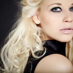 Blondynka z niebieskimi oczami i ciemną oprawą oczu, w czarnej sukience, na czarnym tle.