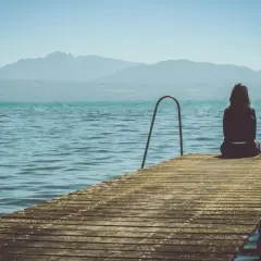 Kobieta siedzi na pomoście, patrząc na spokojne jezioro i góry. To chwila refleksji, być może jeden z etapów rozstania.