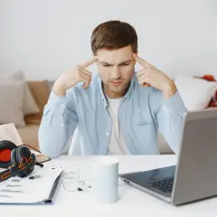 Mężczyzna z palcami przy skroniach, zastanawiający się nad problemem. Obok laptop, książka, słuchawki i kubek. To przykład krytycznego myślenia co to.