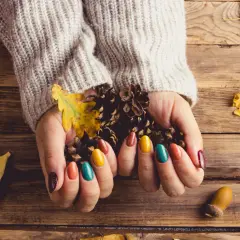 Dłonie w swetrze trzymają szyszki i liście, otoczone jesiennymi dekoracjami. To taki **soft autumn**.