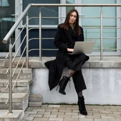 Elegancka kobieta w czarnym płaszczu i spodniach, z laptopem na kolanach, siedzi na schodach. Stylowe czarne płaszcze to podstawa każdej garderoby.
