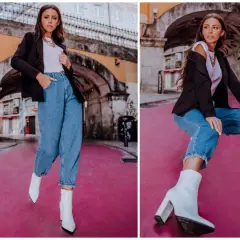 Stylowa kobieta w spodniach mom fit, białych botkach i czarnej marynarce pozuje na tle miejskiej architektury.