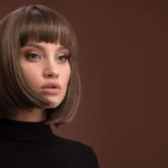Kobieta z modną fryzurą typu bob i grzywką, w czarnym golfie, na brązowym tle.