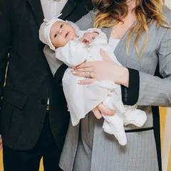 Elegancko ubrani rodzice z dzieckiem w białej szatce. Stylizacja na chrzciny: garnitur dla taty, żakiet dla mamy.