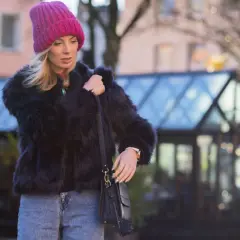Krótkie futerko stylizacje: blondynka w różowej czapce i czarnym futerku, z jeansami i torebką, na tle miasta.