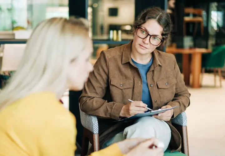 Psycholog a psychoterapeuta w okularach uważnie słucha pacjentki, notując coś na kartce.