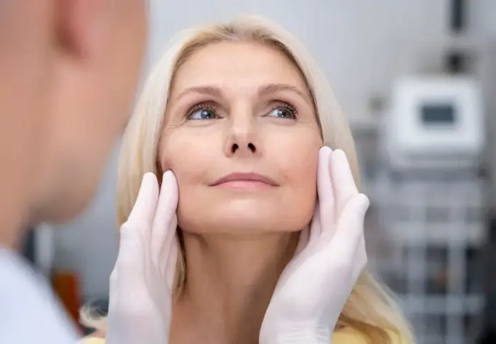 Kobieta z blond włosami poddawana ocenie przed zabiegiem, który może być liftingiem twarzy. Dłonie w rękawiczkach delikatnie dotykają jej policzków.