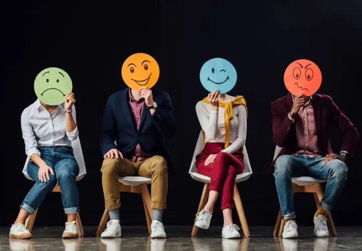 Cztery osoby siedzą na krzesłach, zasłaniając twarze okrągłymi kartkami z różnymi emotikonami. Widać, ile jest emocji: smutek, radość, mrugnięcie i złość.