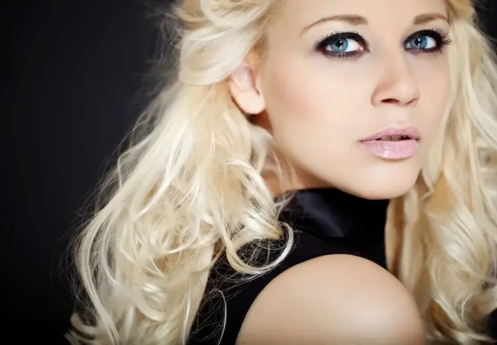 Blondynka z niebieskimi oczami i ciemną oprawą oczu, w czarnej sukience, na czarnym tle.