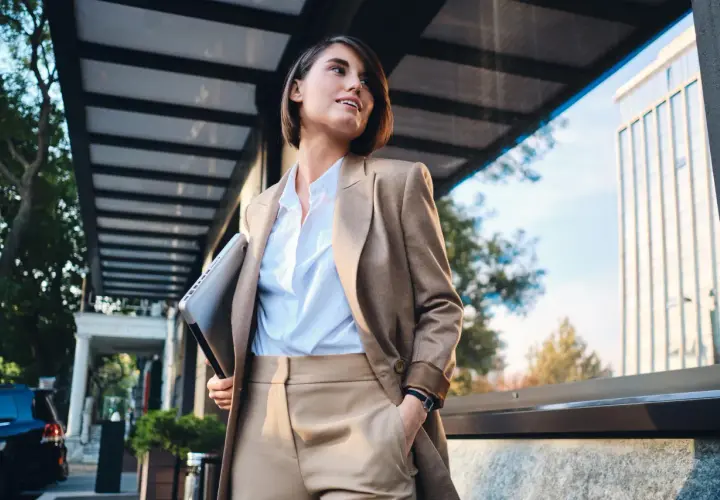 Kobieta w eleganckim stroju, symbolizującym power dressing, trzyma laptopa, gotowa na kolejne wyzwania biznesowe.