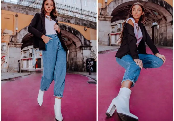 Stylowa kobieta w spodniach mom fit, białych botkach i czarnej marynarce pozuje na tle miejskiej architektury.