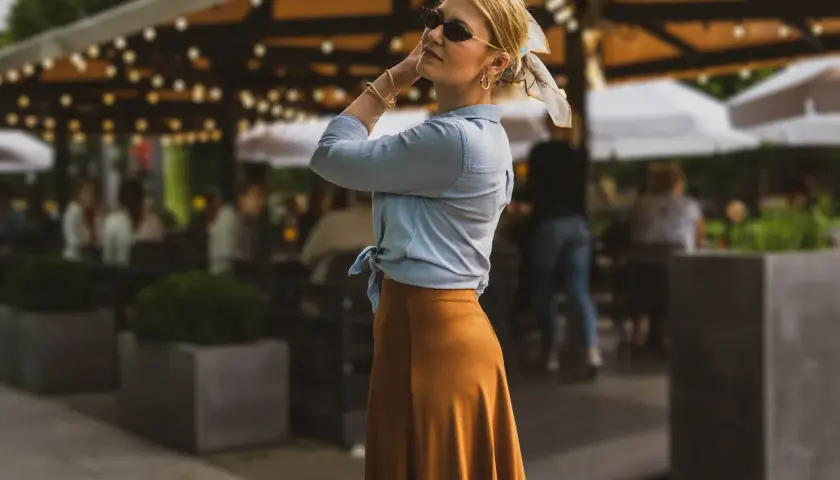 Stylowa kobieta w letniej stylizacji, pokazuje jak zawiązać koszulę. W tle ogródek kawiarniany.