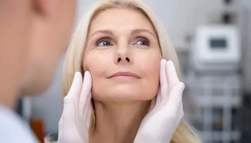 Kobieta z blond włosami poddawana ocenie przed zabiegiem, który może być liftingiem twarzy. Dłonie w rękawiczkach delikatnie dotykają jej policzków.