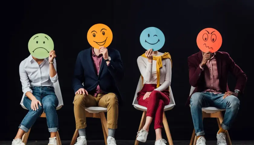 Cztery osoby siedzą na krzesłach, zasłaniając twarze okrągłymi kartkami z różnymi emotikonami. Widać, ile jest emocji: smutek, radość, mrugnięcie i złość.