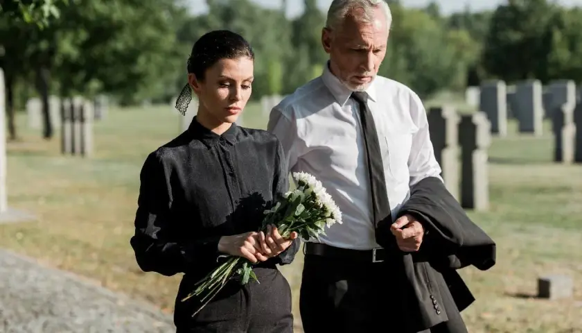 Para pogrzebowa w letni dzień: kobieta w czarnej sukience i mężczyzna w białej koszuli z krawatem, niosący marynarkę i kwiaty.
