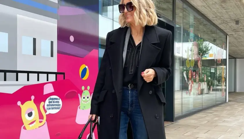 Elegancka stylizacja na imprezę: czarny płaszcz, jeansy i stylowe dodatki. Kobieta z blond włosami i okularami przeciwsłonecznymi pozuje na tle kolorowego muralu z kosmitami.