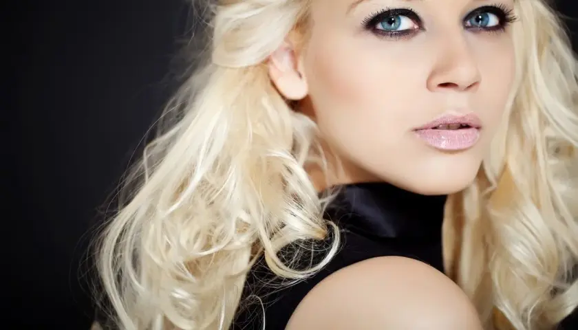 Blondynka z niebieskimi oczami i ciemną oprawą oczu, w czarnej sukience, na czarnym tle.