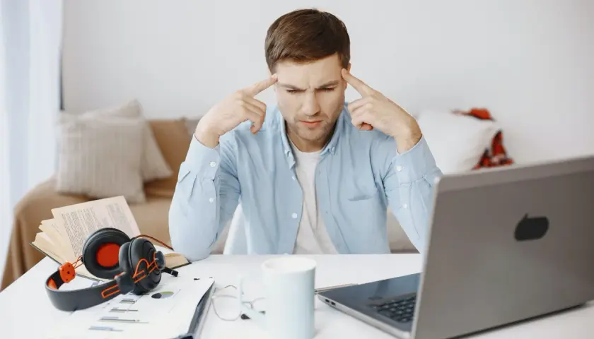 Mężczyzna z palcami przy skroniach, zastanawiający się nad problemem. Obok laptop, książka, słuchawki i kubek. To przykład krytycznego myślenia co to.