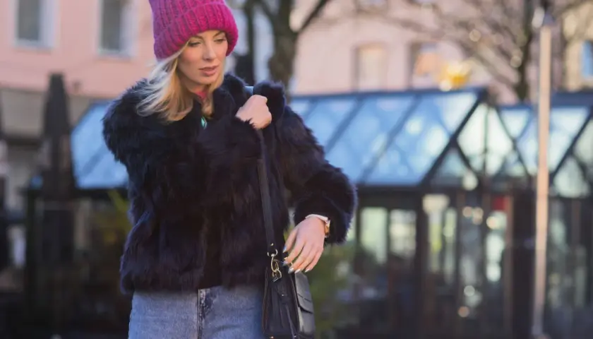 Krótkie futerko stylizacje: blondynka w różowej czapce i czarnym futerku, z jeansami i torebką, na tle miasta.