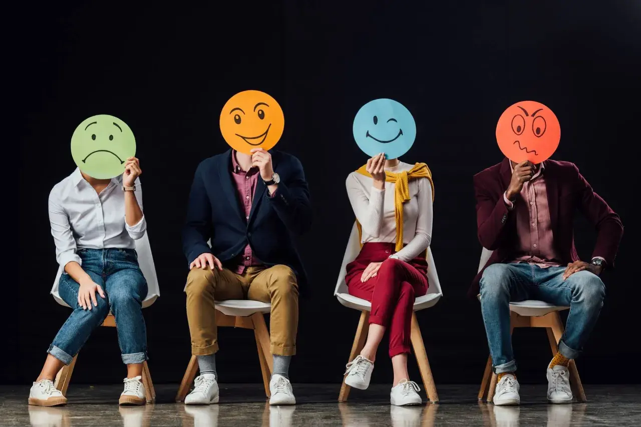 Cztery osoby siedzą na krzesłach, zasłaniając twarze okrągłymi kartkami z różnymi emotikonami. Widać, ile jest emocji: smutek, radość, mrugnięcie i złość.