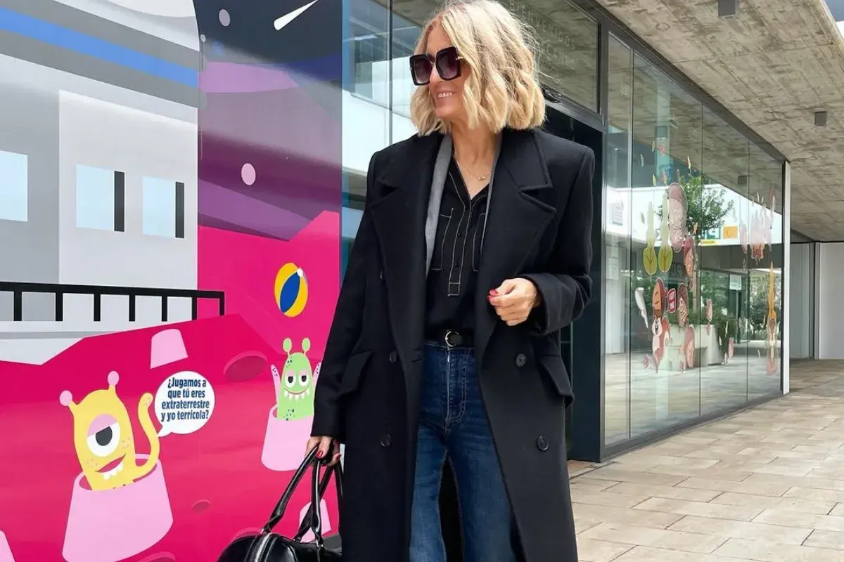 Elegancka stylizacja na imprezę: czarny płaszcz, jeansy i stylowe dodatki. Kobieta z blond włosami i okularami przeciwsłonecznymi pozuje na tle kolorowego muralu z kosmitami.