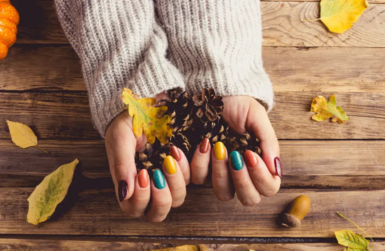 Dłonie w swetrze trzymają szyszki i liście, otoczone jesiennymi dekoracjami. To taki **soft autumn**.