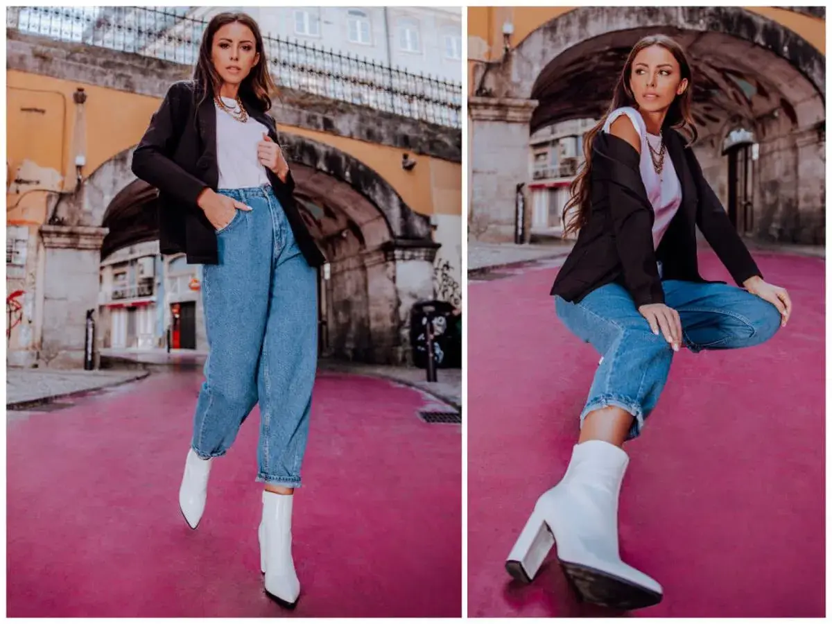 Stylowa kobieta w spodniach mom fit, białych botkach i czarnej marynarce pozuje na tle miejskiej architektury.