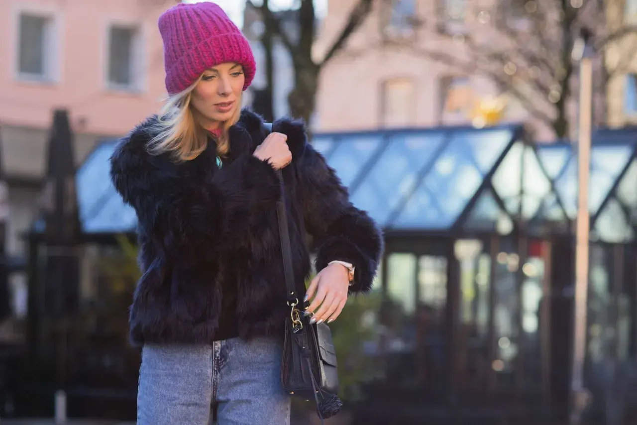 Krótkie futerko stylizacje: blondynka w różowej czapce i czarnym futerku, z jeansami i torebką, na tle miasta.