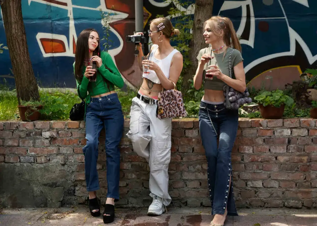 Trzy dziewczyny w bojówkach i crop topach pozują przy murze z graffiti. Jedna z nich trzyma aparat. Jakie buty do bojówek?