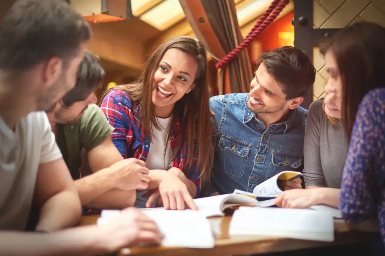 Grupa studentów dyskutuje, szukając odpowiedzi na pytania otwarte. Przykładem jest dziewczyna wskazująca palcem na książkę.