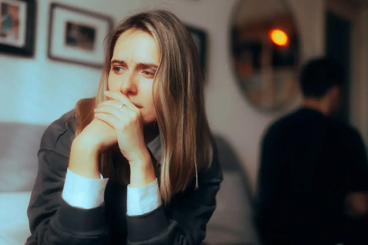 Kobieta zamyślona, z rękami przy twarzy, siedzi obok niewyraźnej postaci. Widać między nimi dystans emocjonalny.