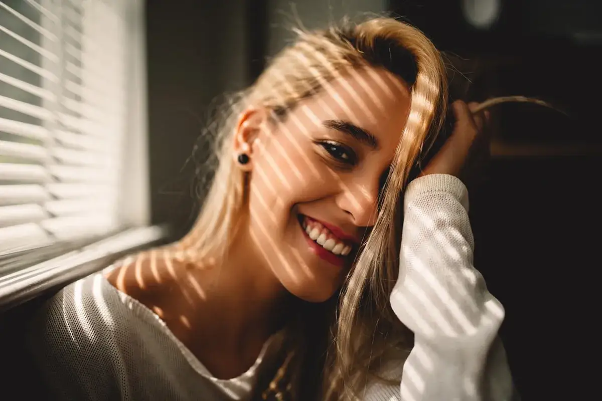 Uśmiechnięta blondynka z pozytywnymi cechami charakteru, oświetlona promieniami słońca przez żaluzje.