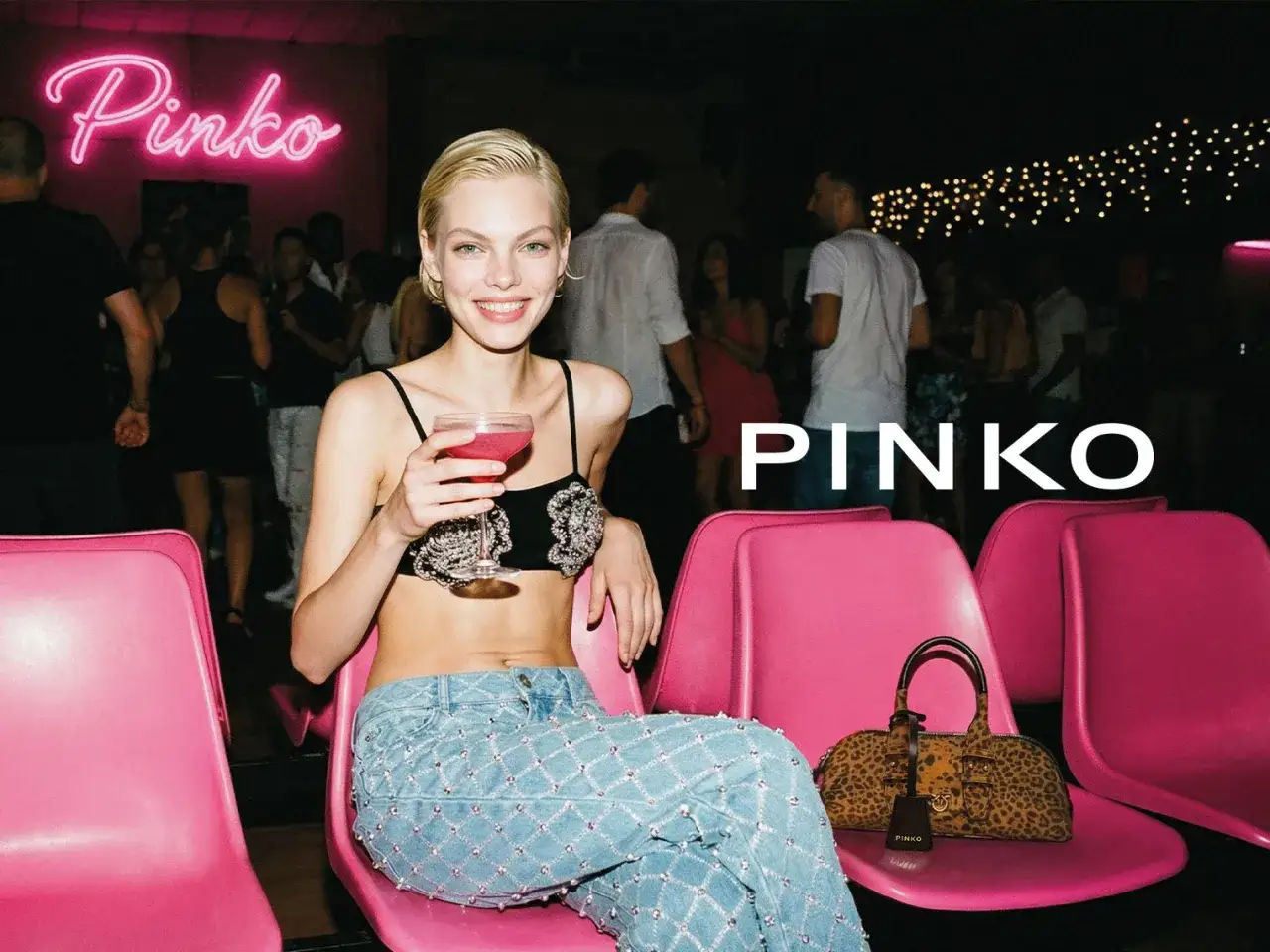 Modelka w jeansach i ozdobnym topie, z drinkiem w ręku, siedzi na różowym krześle. Obok torebka Pinko. Idealne torebka Pinko stylizacje na wieczór.