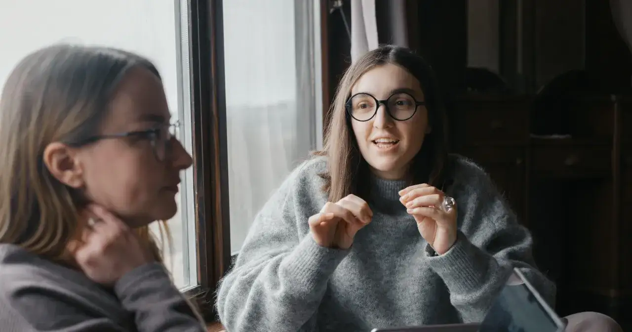 Dwie kobiety rozmawiają przy oknie. Jedna zadaje pytania drugiej połówce, gestykulując.